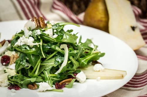 Arugula salad with crumbled cheese, dried cranberries and pecans beside a wedge of cheese