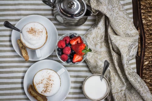 Two cappuccinos, a bowl of berries and a pot of cream on striped linen